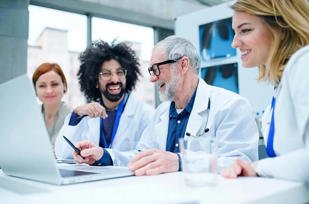 Smiling adults wearing white coats in a lab are seated in front of a laptop. This post looks at why people hesitate to participate in clinical trials. Clinical studies blog.